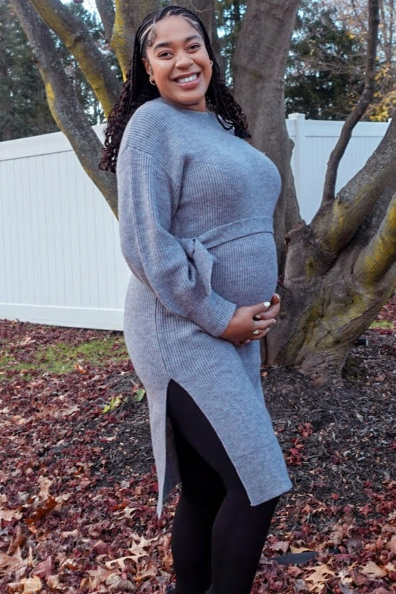 woman wearing a gray knitted maternity dress with side slits, smiling and showing her baby bump outdoors