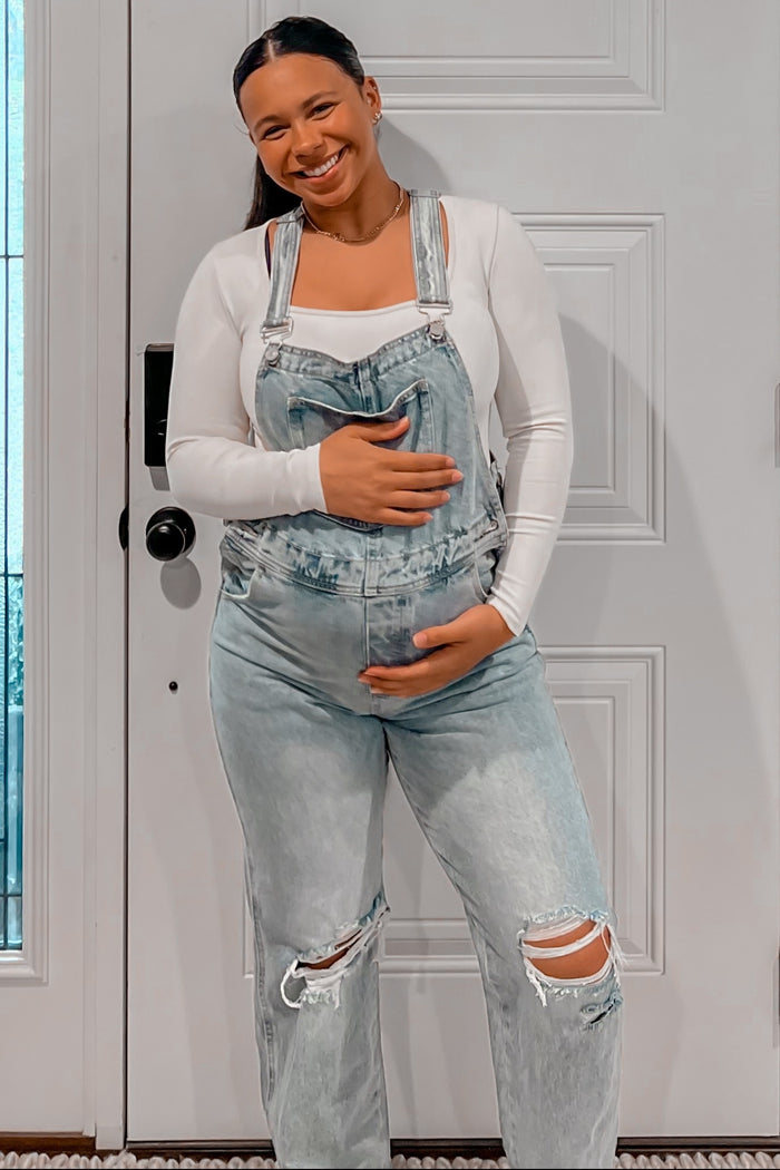 woman wearing a white long sleeve maternity top and distressed denim overalls, holding her baby bump
