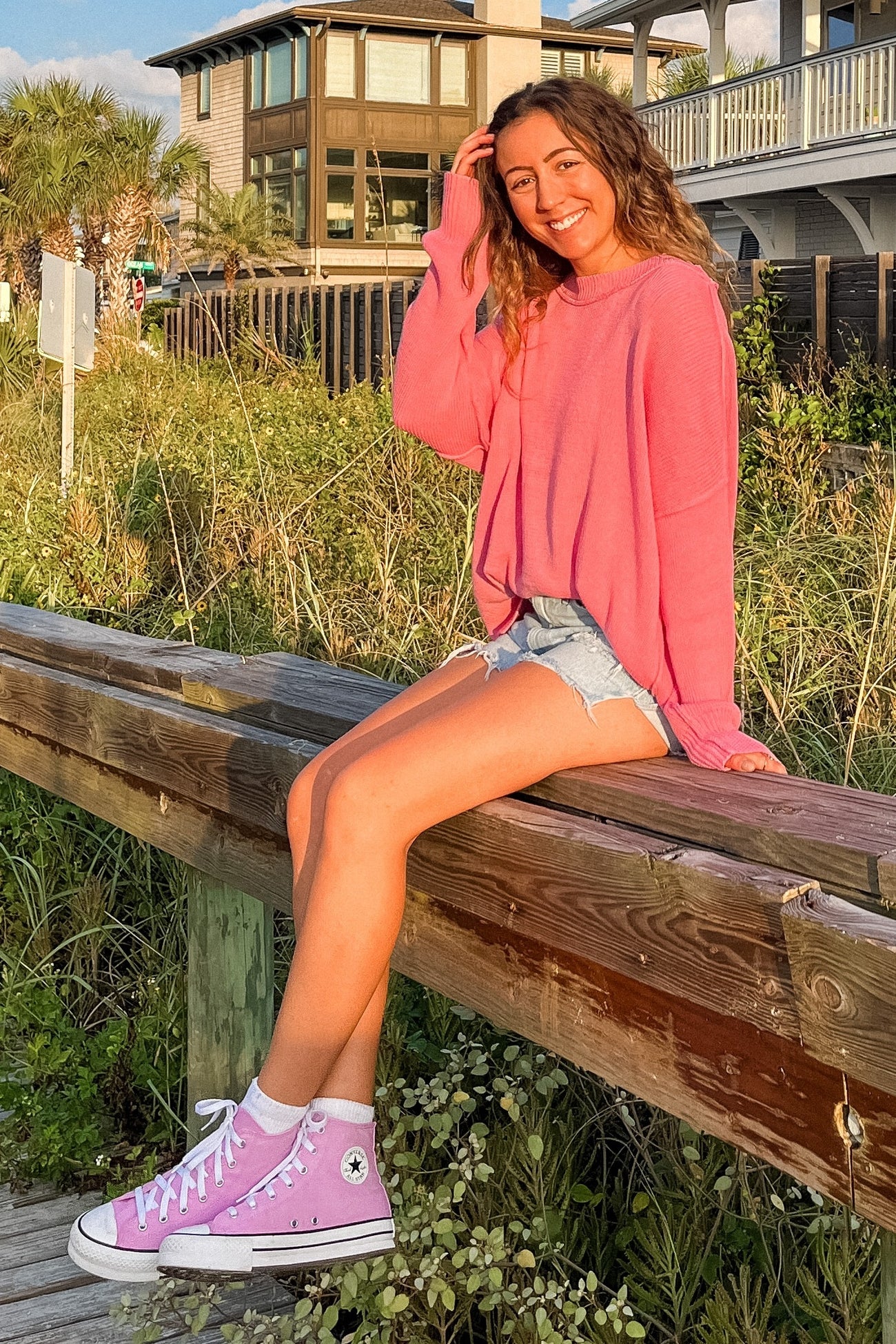 woman wearing a bright pink oversized sweater and denim shorts, sitting on a wooden railing by the beach