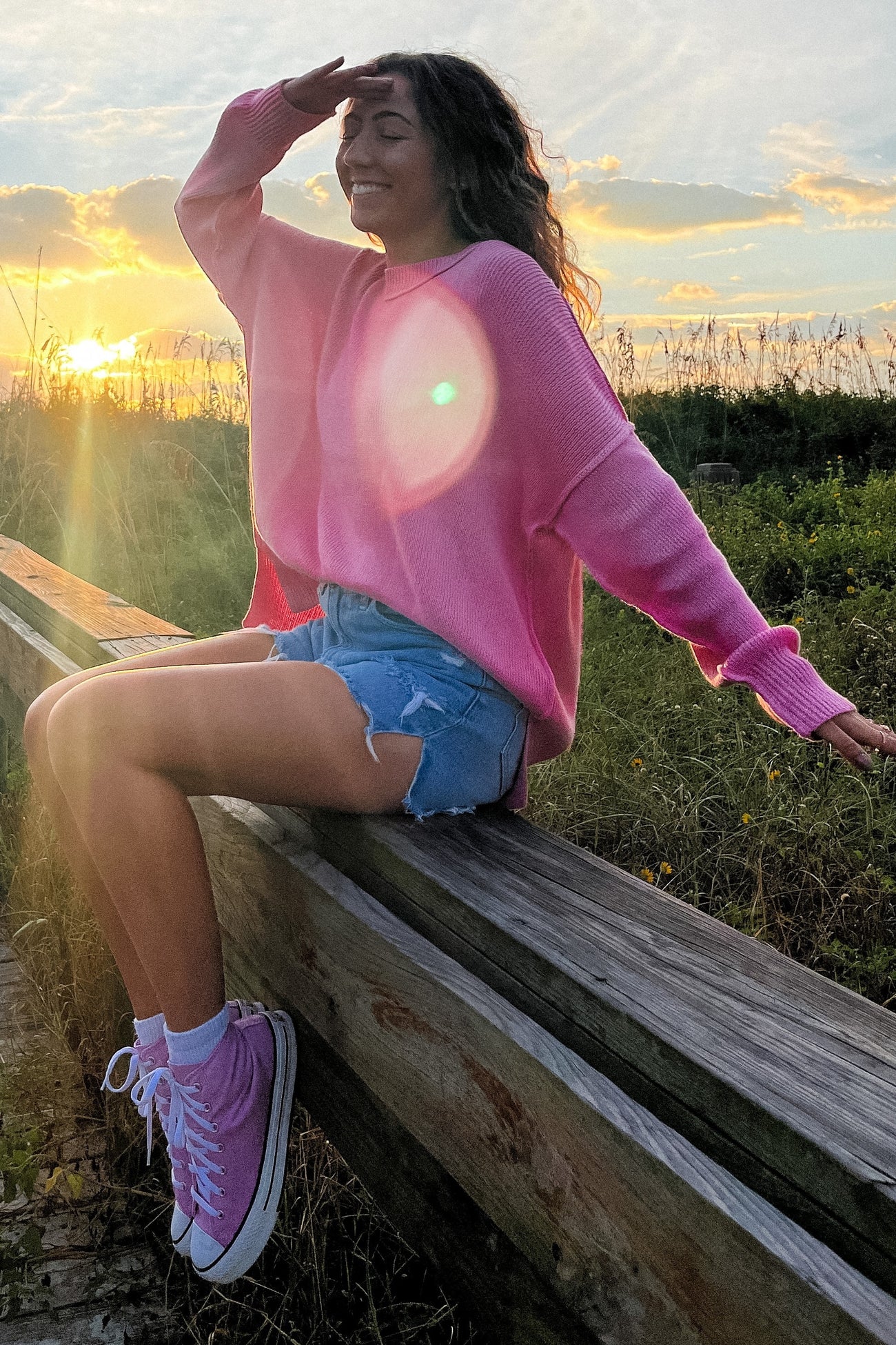 woman wearing a pink oversized sweater, distressed denim shorts, and pink sneakers, sitting on a wooden railing at sunset
