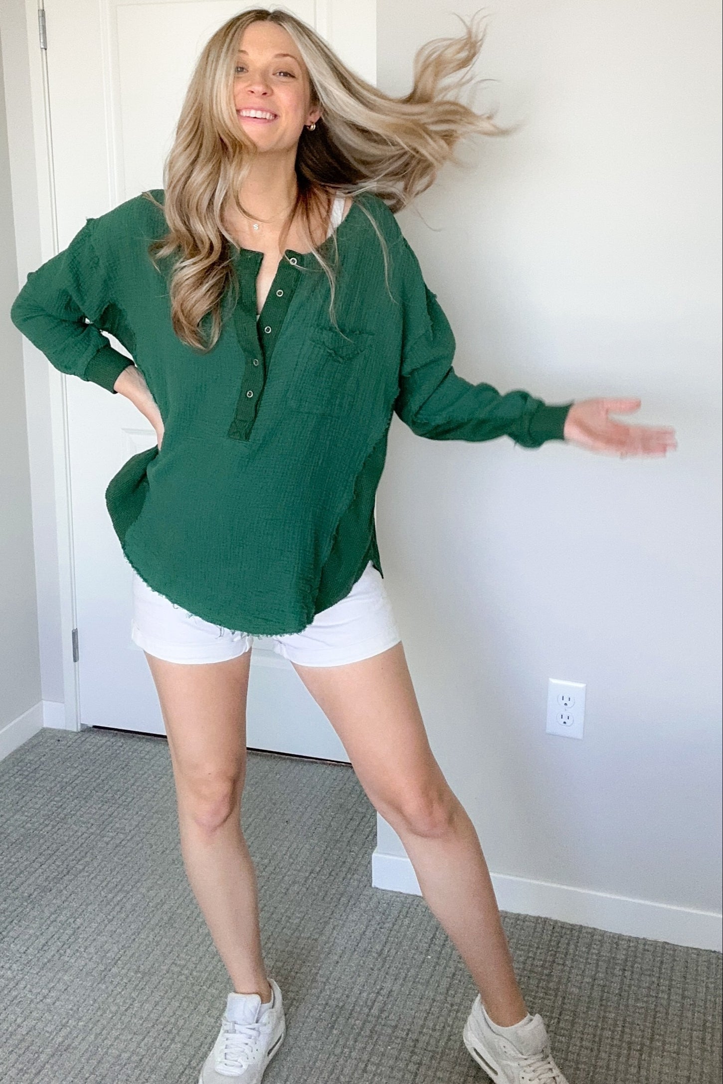 woman wearing a green button-up top and white shorts, smiling with one leg posed and hair flowing