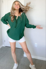 woman wearing a green button-up top and white shorts, smiling with one leg posed and hair flowing