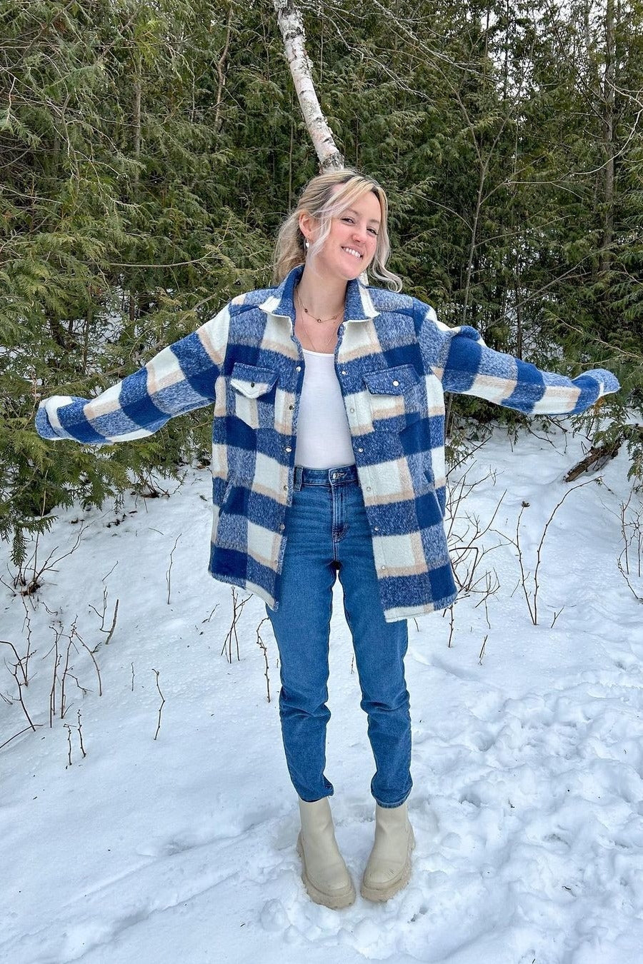 woman wearing a blue and white plaid shacket with a white top, light blue jeans, and cream-colored boots in a snowy setting