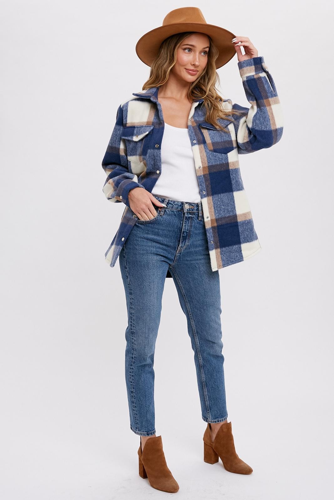 woman wearing a blue and cream plaid shacket with a white tank top, light wash jeans, and brown ankle boots, accessorized with a hat