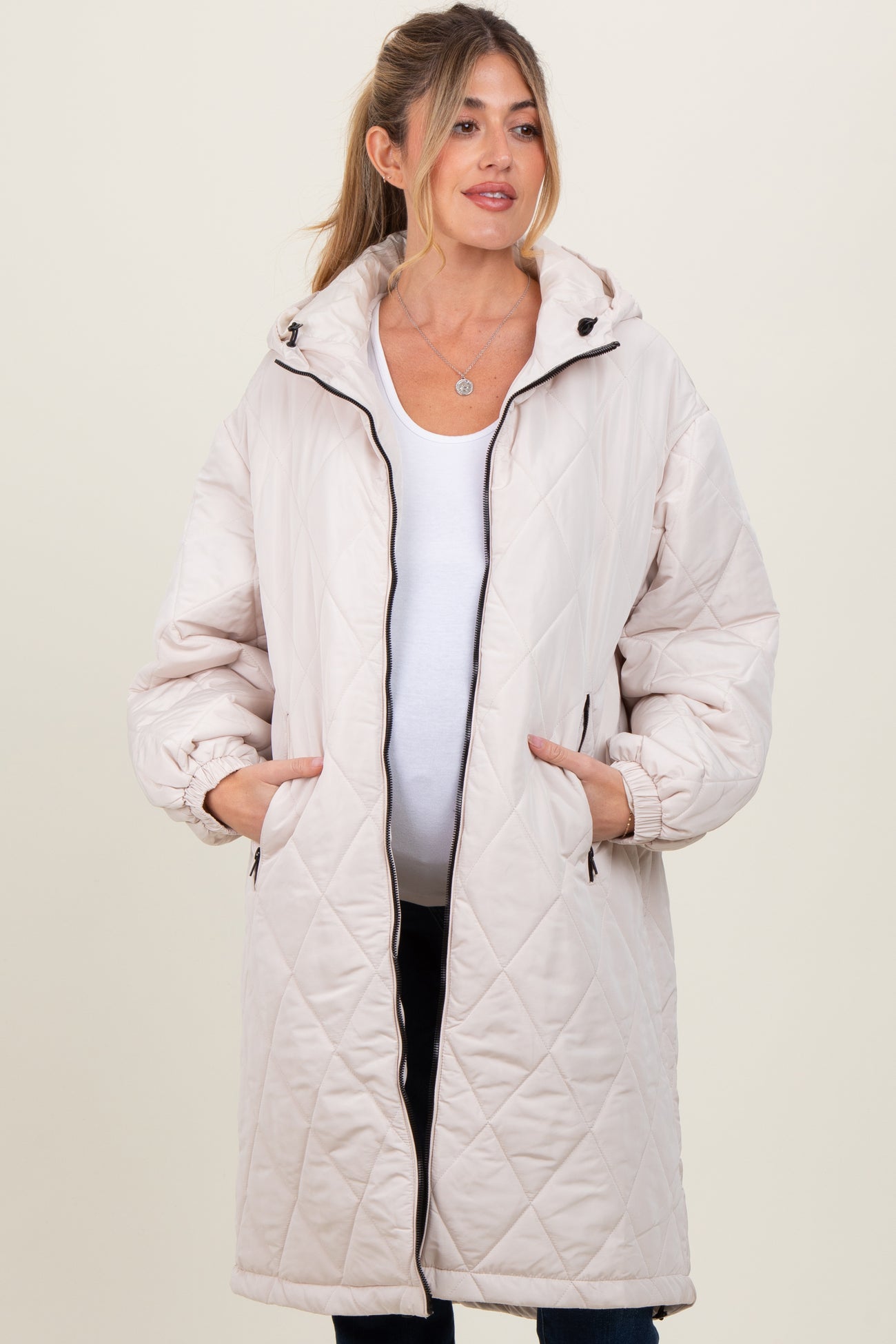 woman wearing a light beige quilted maternity coat with a hood and pockets, baby bump visible