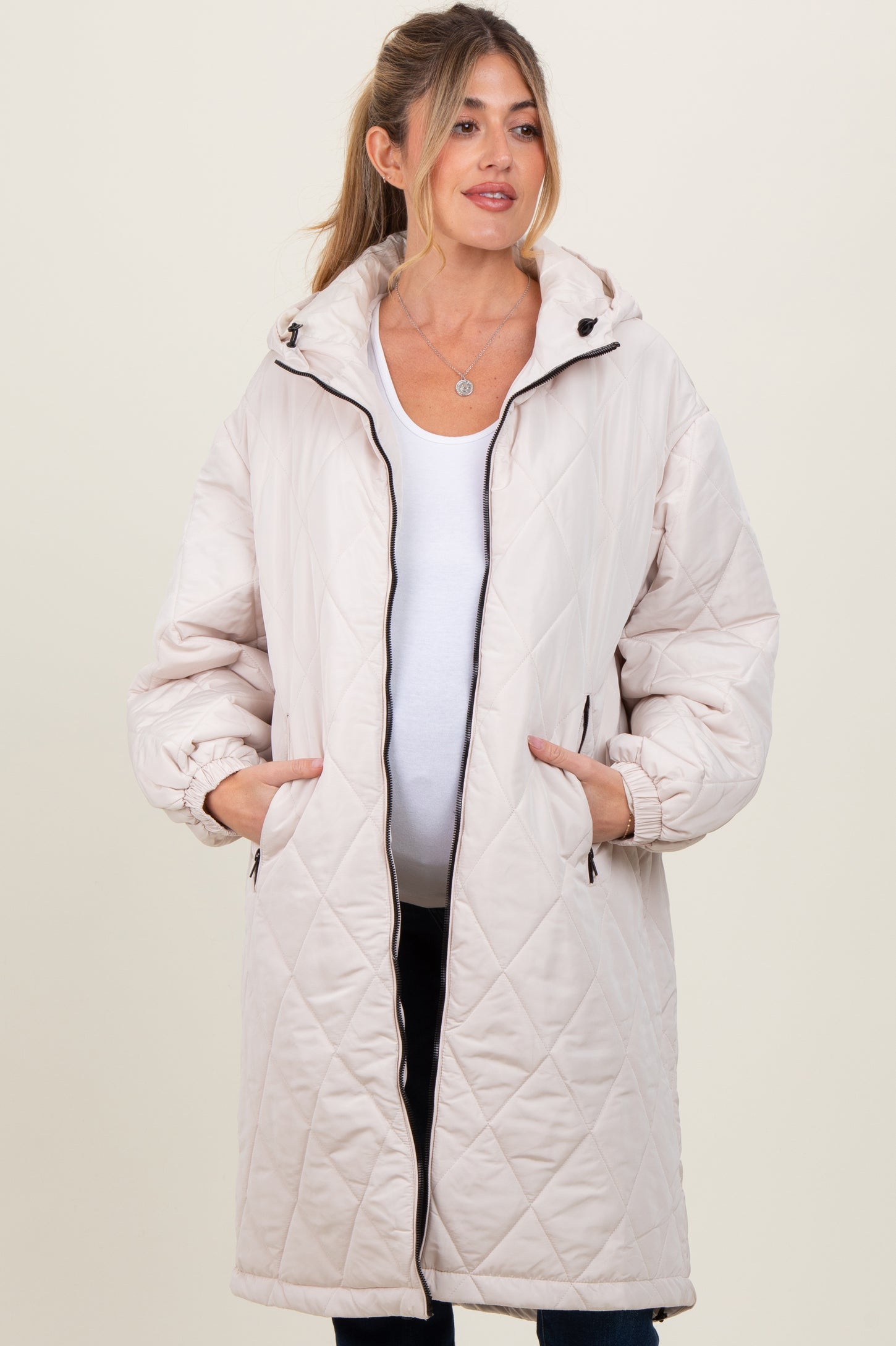 woman wearing a light beige quilted maternity coat with a hood and pockets, baby bump visible