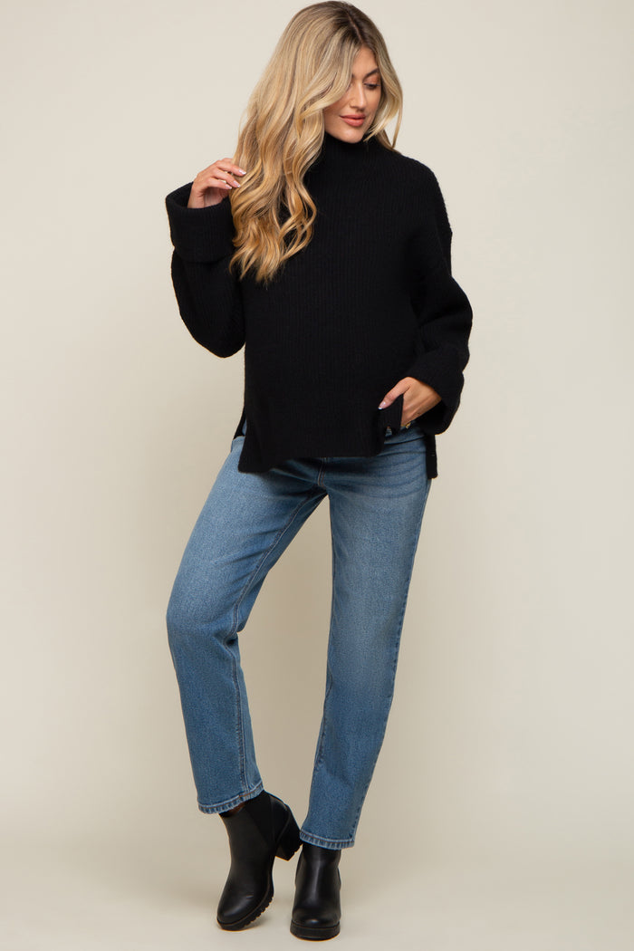 woman wearing a black oversized maternity sweater with side slits and blue straight-leg jeans, posing with one hand on her hip