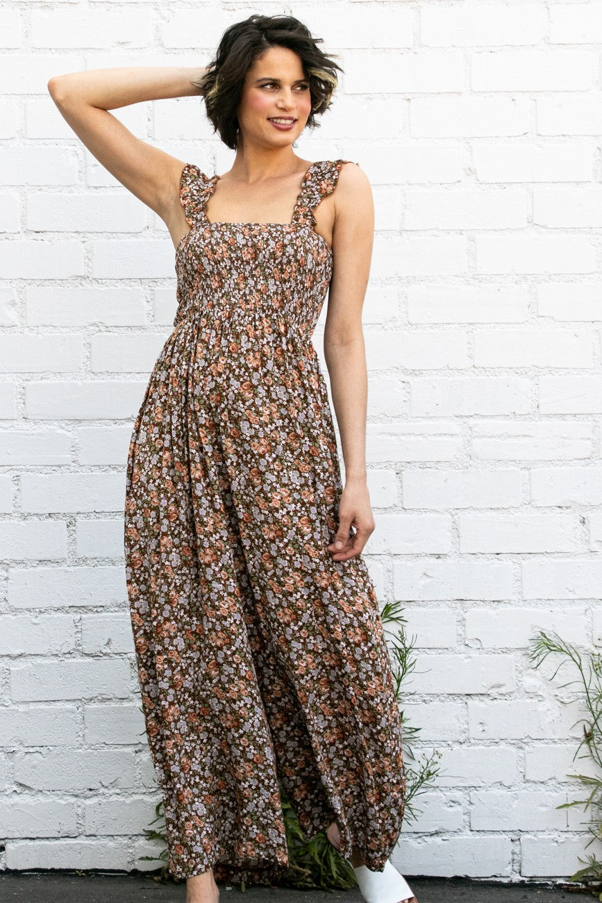 woman wearing a brown floral maxi dress with smocked bodice and ruffled straps, posing against a white brick wall