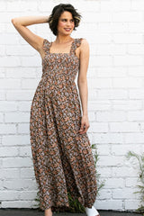 woman wearing a brown floral maxi dress with smocked bodice and ruffled straps, posing against a white brick wall