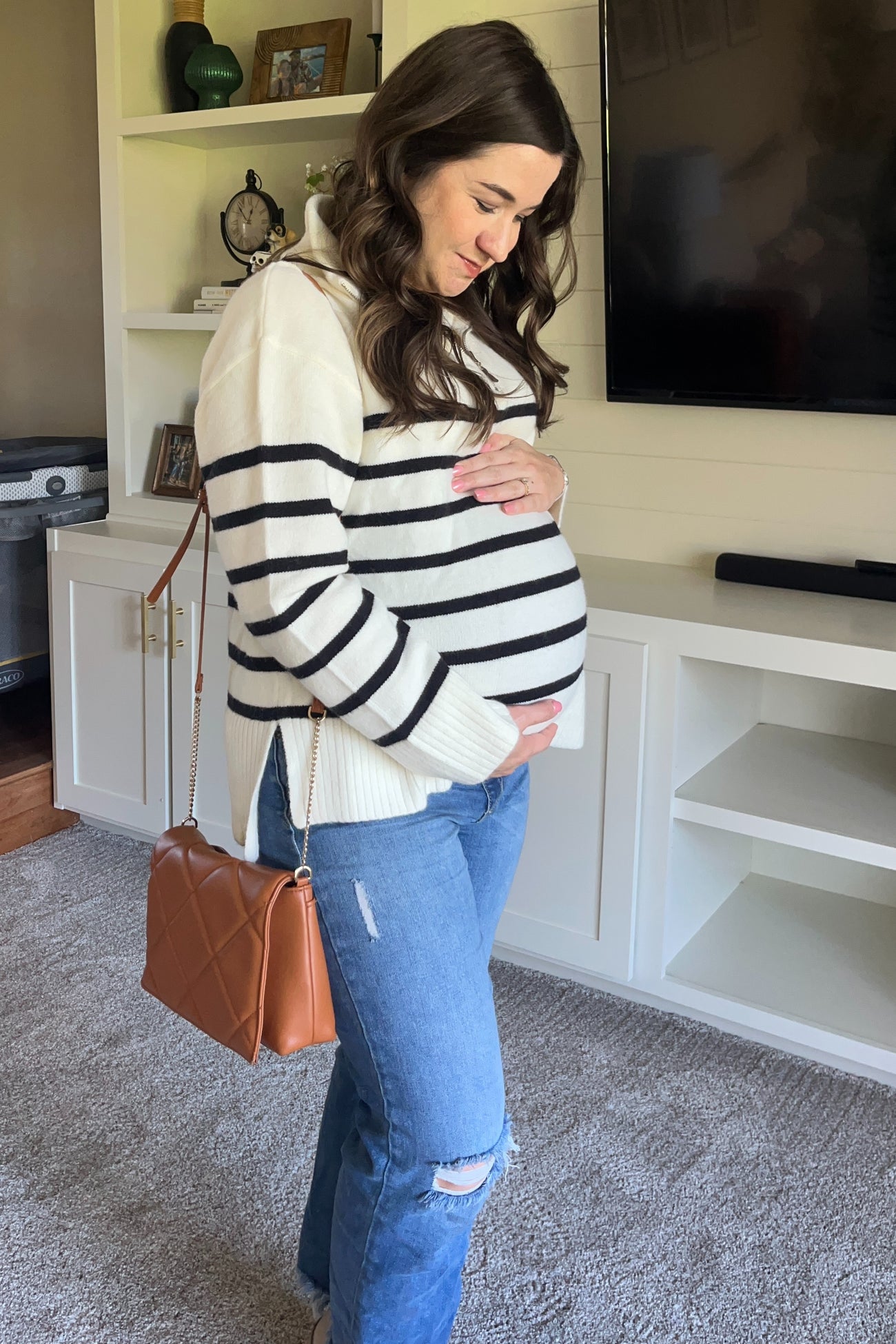 woman wearing a cream and black striped maternity sweater and distressed jeans, holding her baby bump