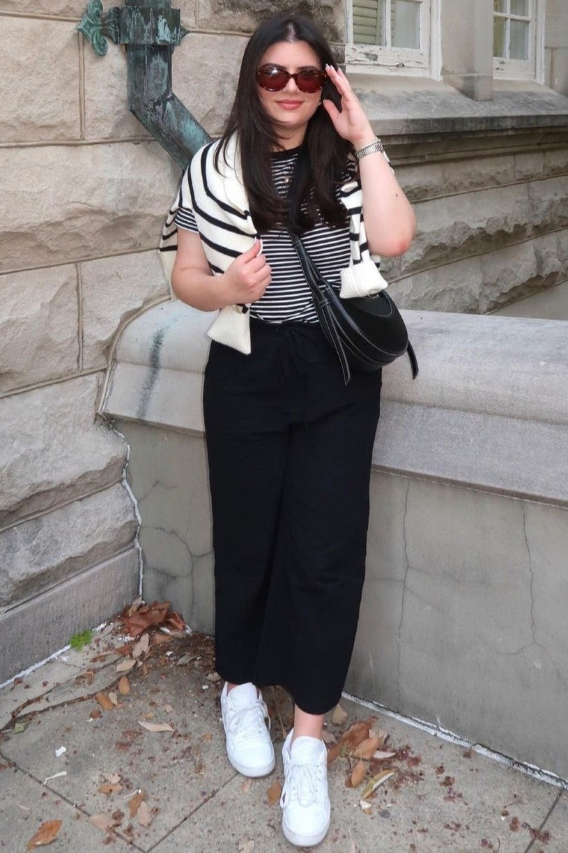 woman wearing a striped black and white top with a black skirt, white sneakers, and sunglasses, standing by a stone wall