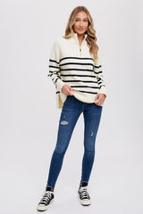 woman wearing a cream and black striped zip-up sweater with side slits and blue skinny jeans, standing and smiling