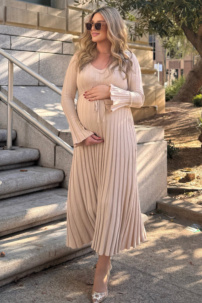 woman wearing a beige pleated maternity dress and holding her baby bump while standing outdoors