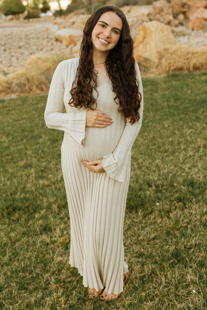woman wearing a cream long sleeve maternity dress with pleats and holding her baby bump outdoors
