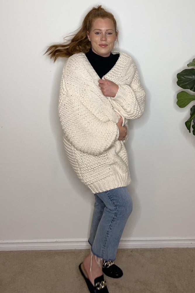 woman wearing a chunky cream oversized sweater with rolled sleeves and light wash cropped jeans, smiling against a plain background