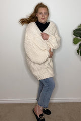 woman wearing a chunky cream oversized sweater with rolled sleeves and light wash cropped jeans, smiling against a plain background