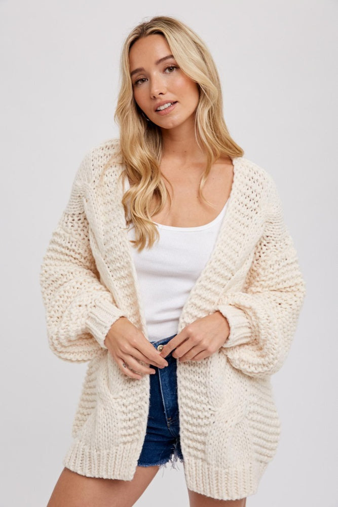 woman wearing a cream oversized cable knit cardigan over a white tank top and blue denim shorts