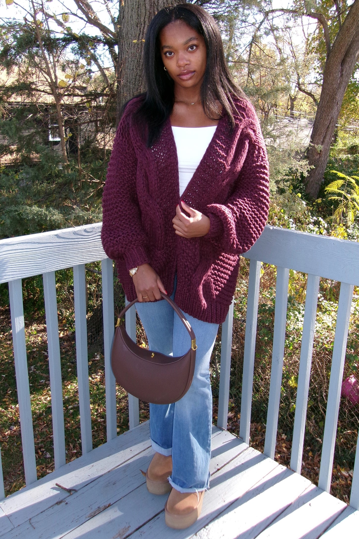woman wearing a burgundy chunky knit cardigan, white top, and light blue flared jeans, holding a brown handbag