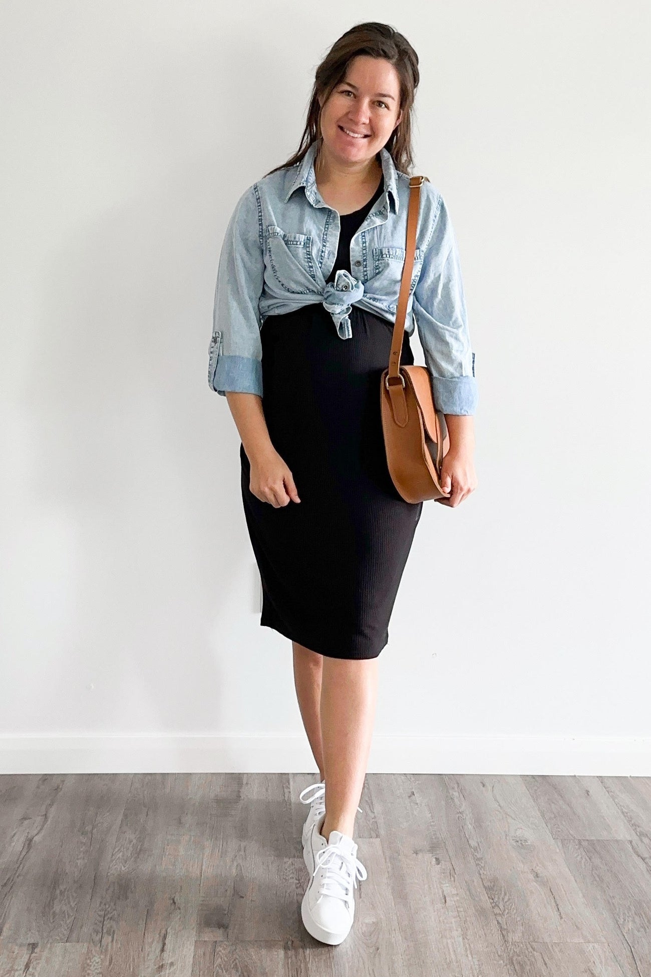 woman wearing a black maternity dress with a knotted denim shirt tied around her waist and white sneakers, smiling and holding a brown bag