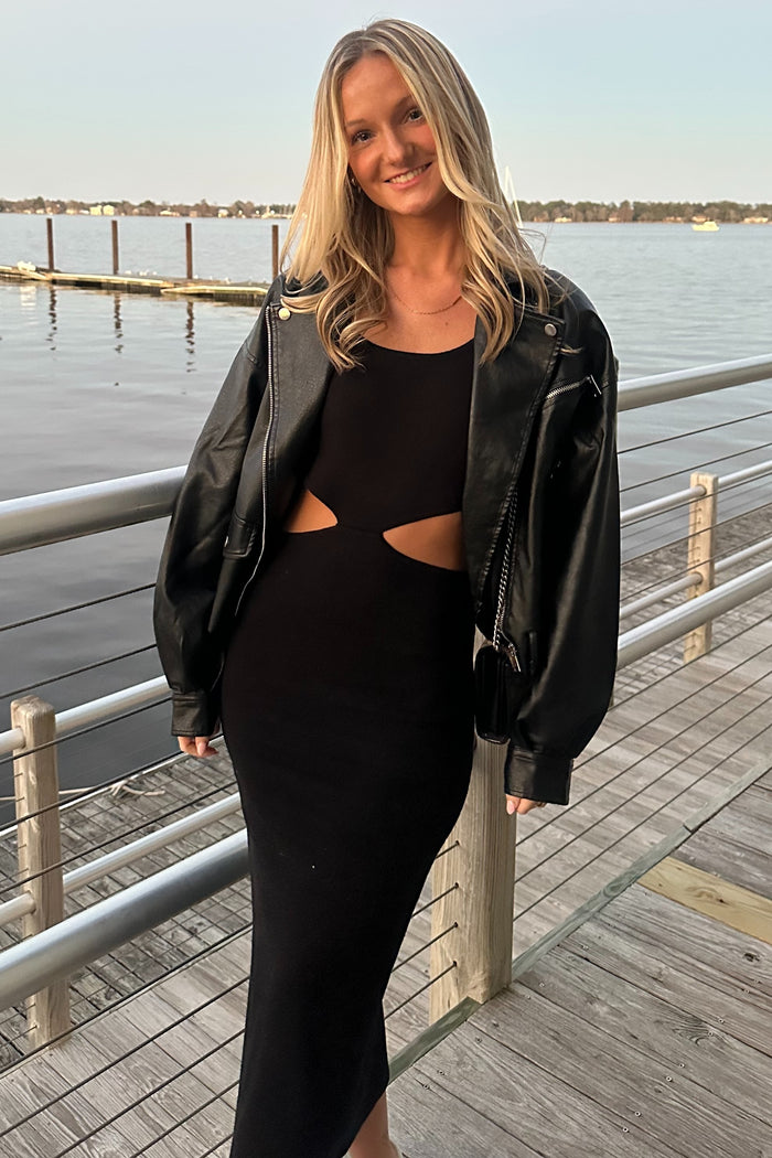 woman wearing a black cut-out midi dress with a black leather jacket, standing by the water at sunset