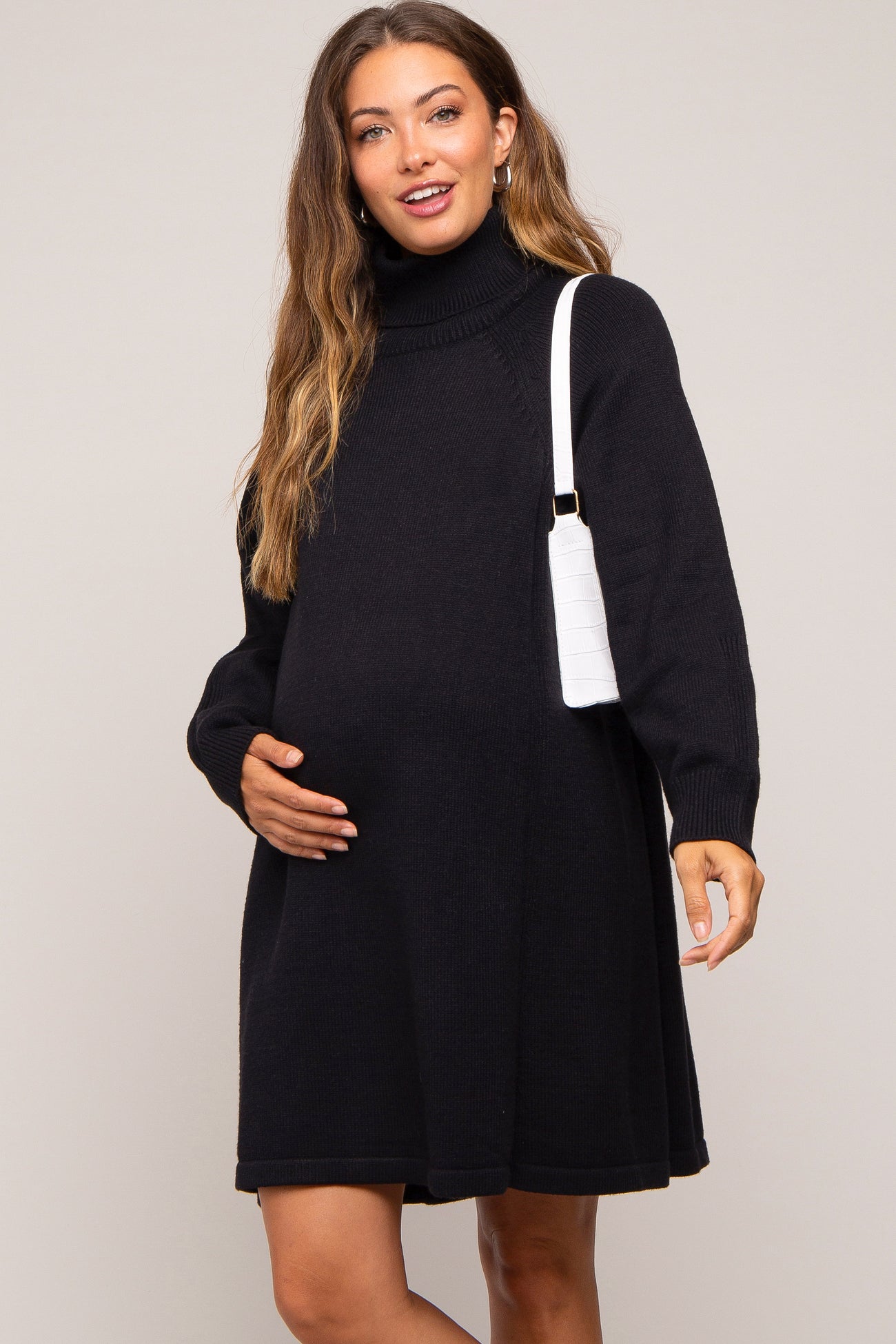 woman wearing a black turtleneck maternity dress and holding her baby bump with a white handbag over her shoulder