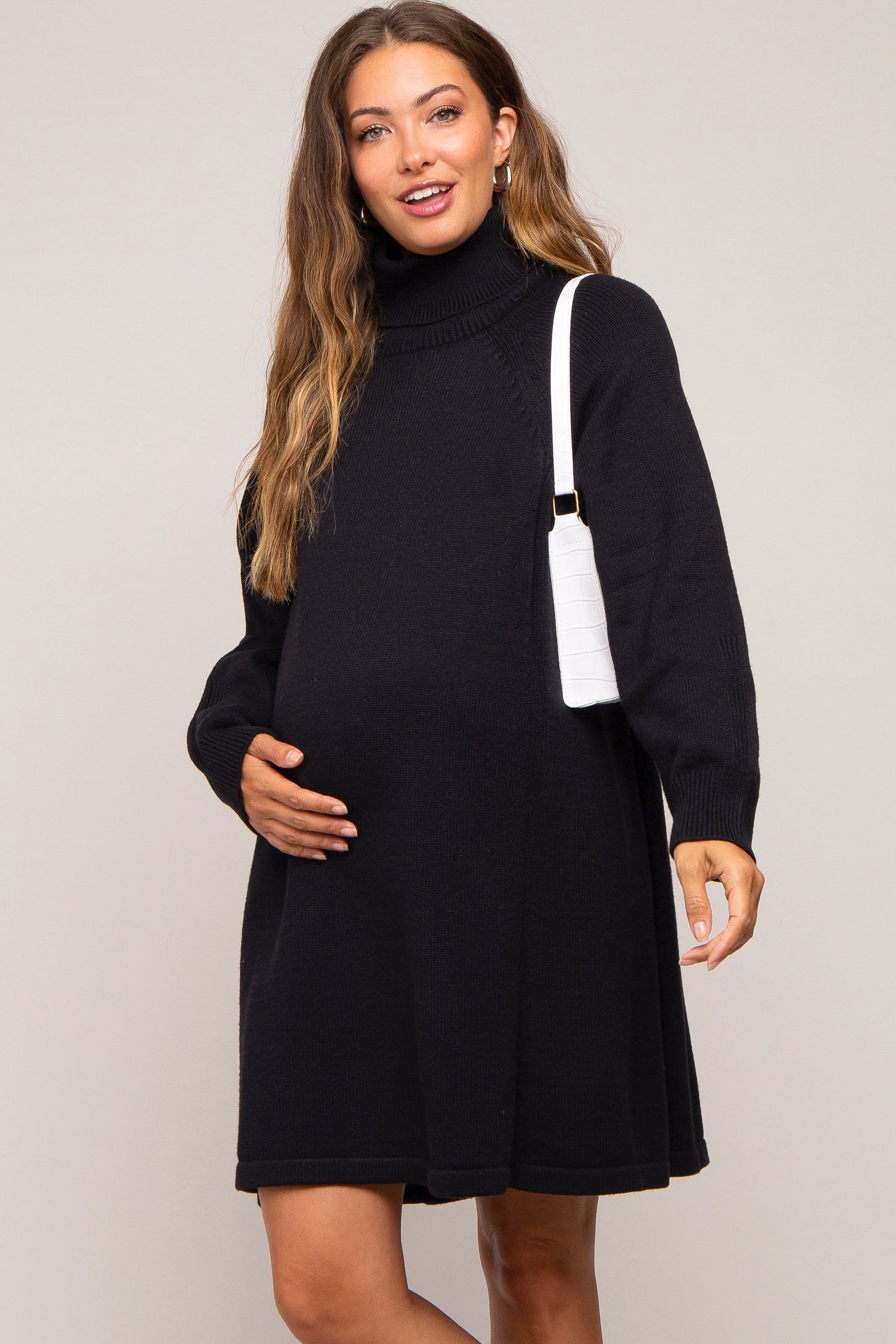 woman wearing a black turtleneck maternity dress and holding her baby bump with a white handbag over her shoulder