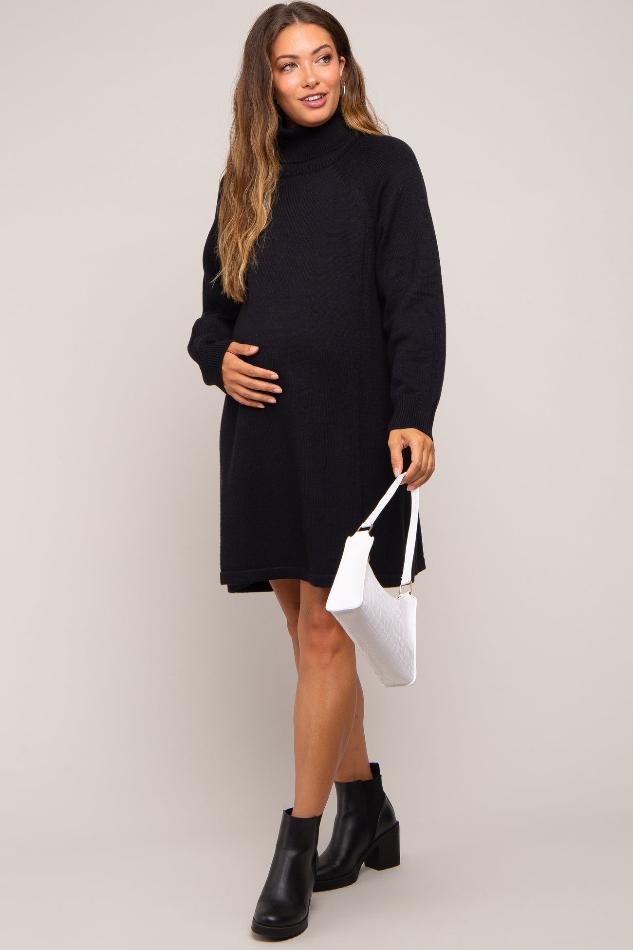 woman wearing a black knit maternity sweater dress and holding her baby bump with a white handbag
