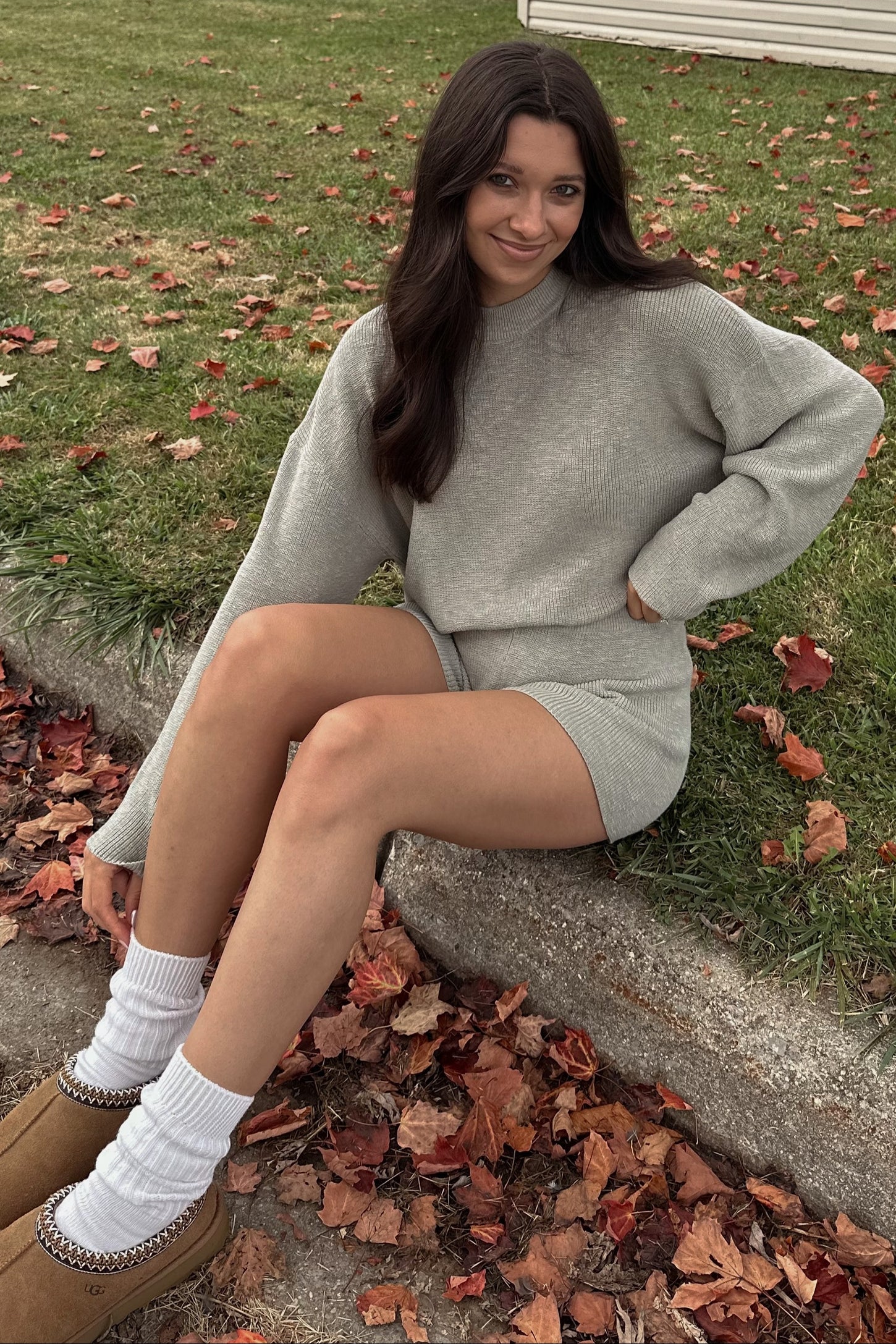 woman wearing a light gray oversized sweater and matching shorts sitting on the ground surrounded by autumn leaves