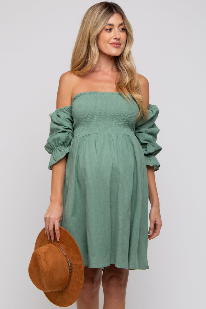 woman wearing a green off-the-shoulder maternity dress with puff sleeves and holding a brown hat, showing a baby bump
