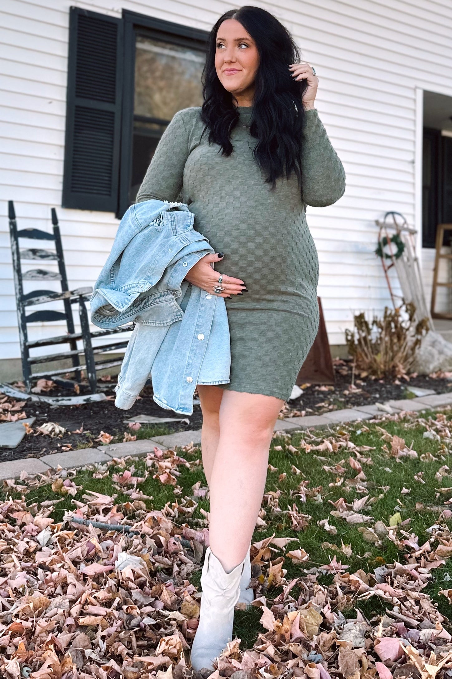 woman wearing a green knitted maternity dress and holding her baby bump, with a denim jacket draped over her arm
