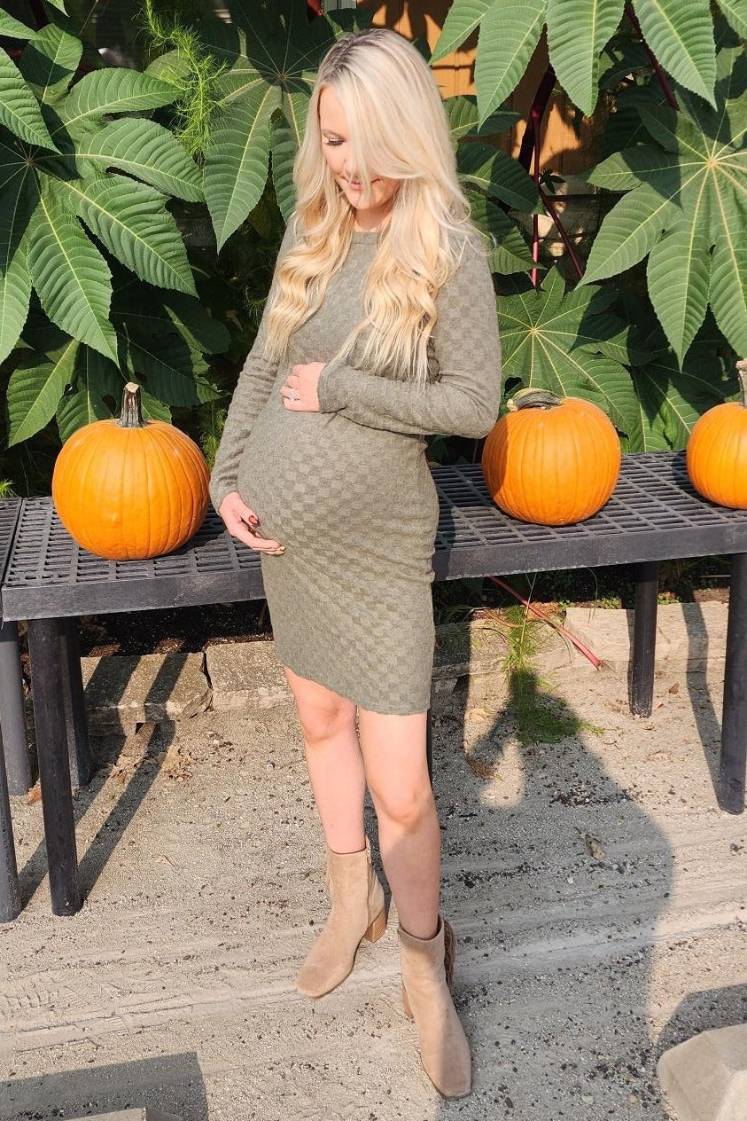 woman wearing a dark green maternity dress with a baby bump, standing beside pumpkins in a garden setting