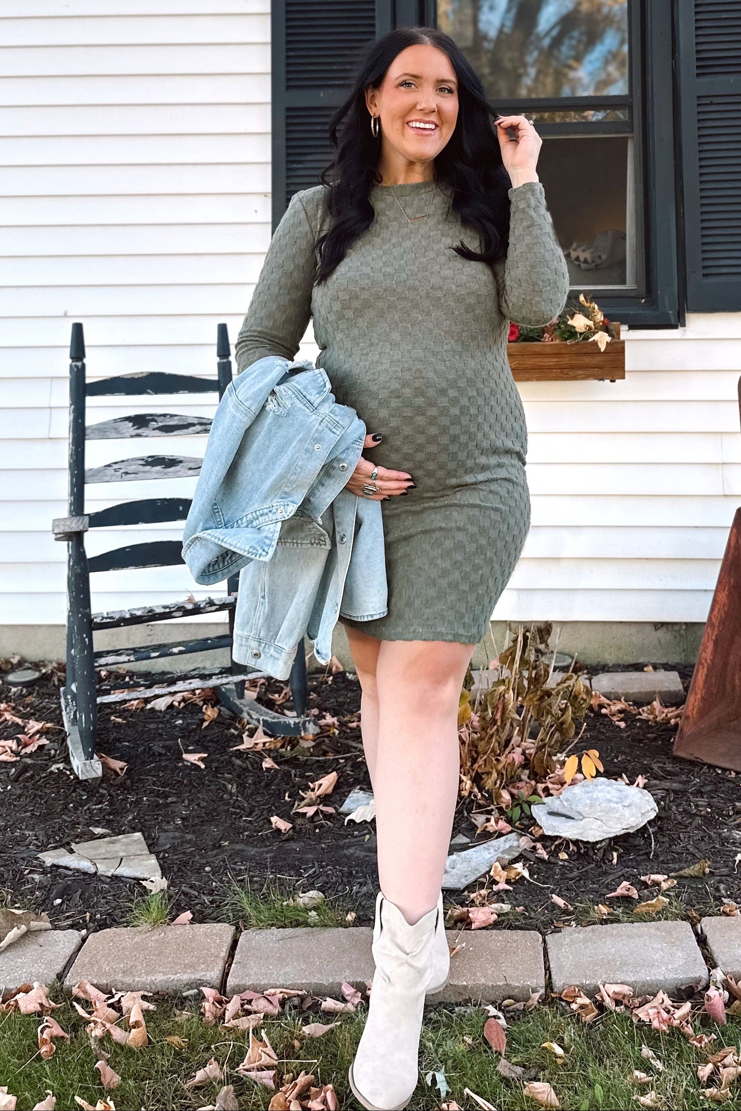 woman wearing a green long sleeve maternity dress and holding her baby bump, with a denim jacket and white boots