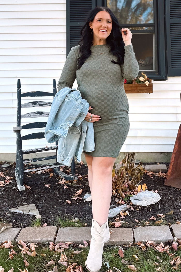 woman wearing a green long sleeve maternity dress and holding her baby bump, with a denim jacket and white boots