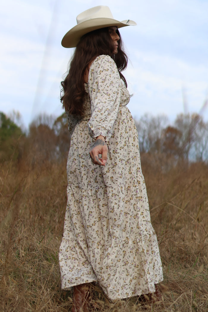 woman wearing a floral print maternity maxi dress and a wide-brimmed hat, standing in a field with a visible baby bump