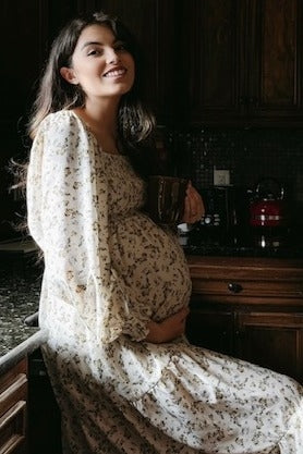 woman wearing a cream floral maternity dress and holding a cup, showing her baby bump while sitting in the kitchen