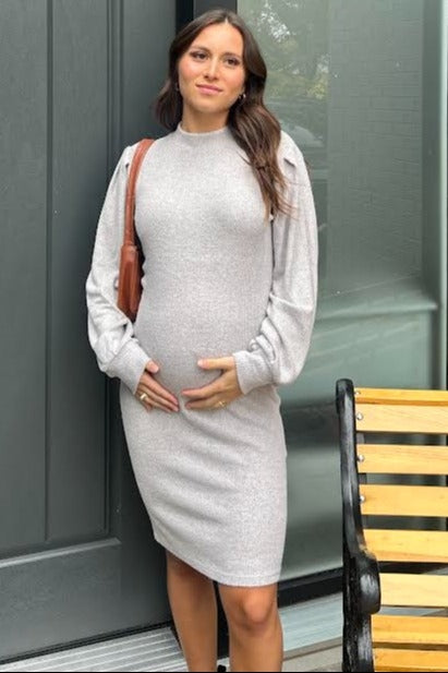 woman wearing a light gray maternity dress with balloon sleeves and a baby bump, standing near a doorway