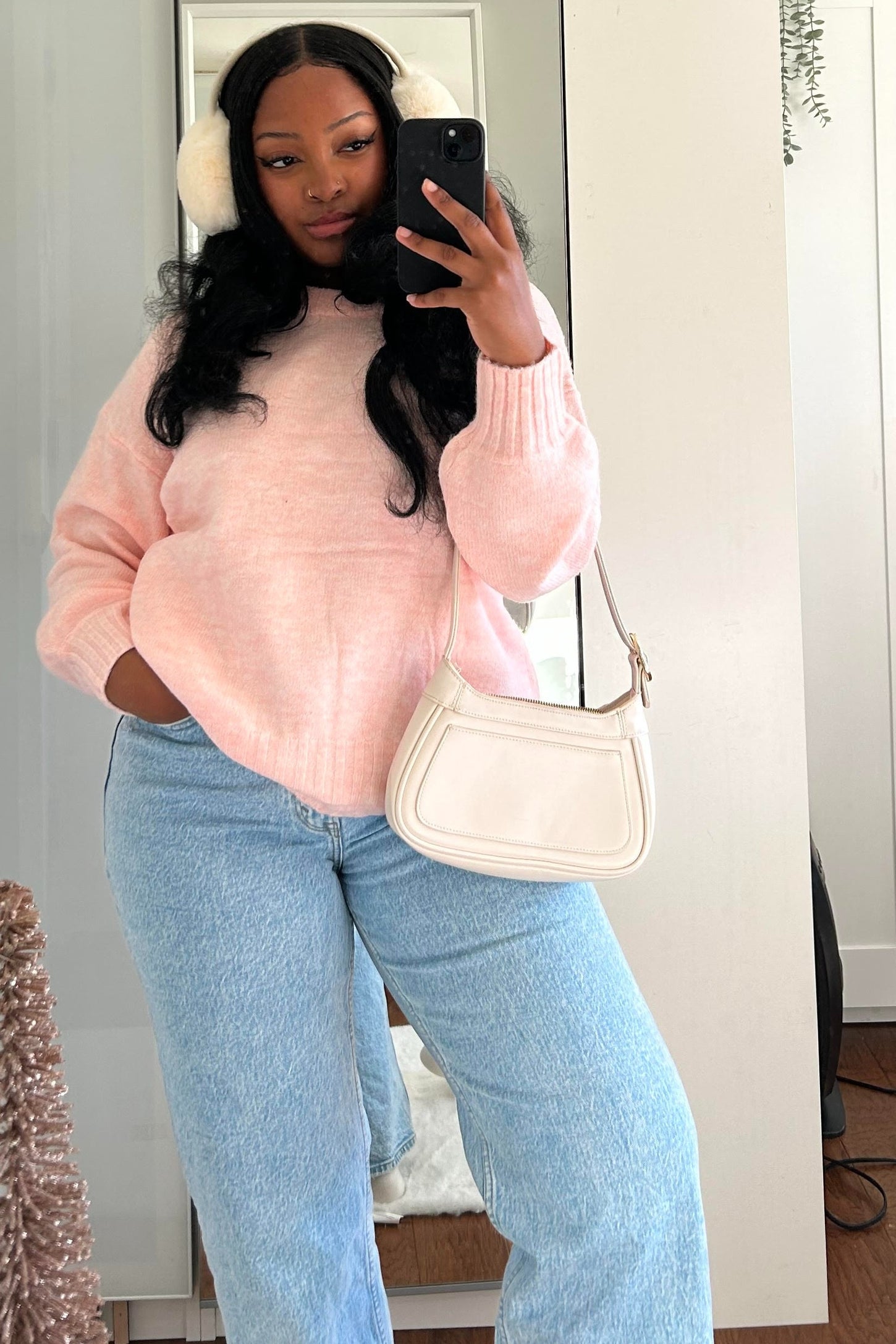 woman wearing a soft pink oversized sweater and light blue wide-leg jeans, holding a cream crossbody bag while taking a mirror selfie