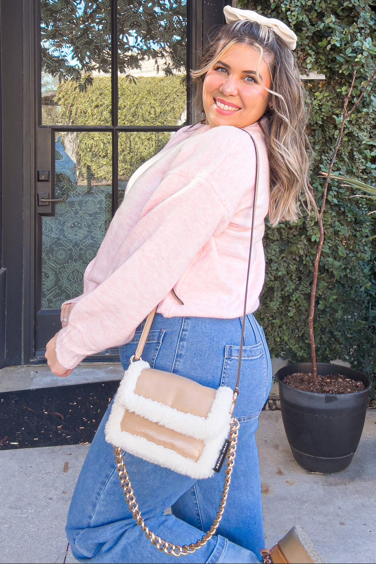woman wearing a pink long sleeve sweater and blue high-waisted jeans, holding a cream-colored shoulder bag with a chain strap