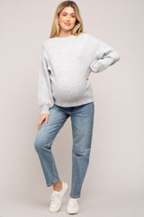 woman wearing a light gray maternity sweater and blue jeans, showing her baby bump, standing in a relaxed pose
