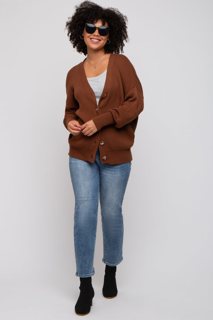 woman wearing a brown oversized cardigan with buttons, gray top, blue jeans, and black ankle boots, smiling and wearing sunglasses