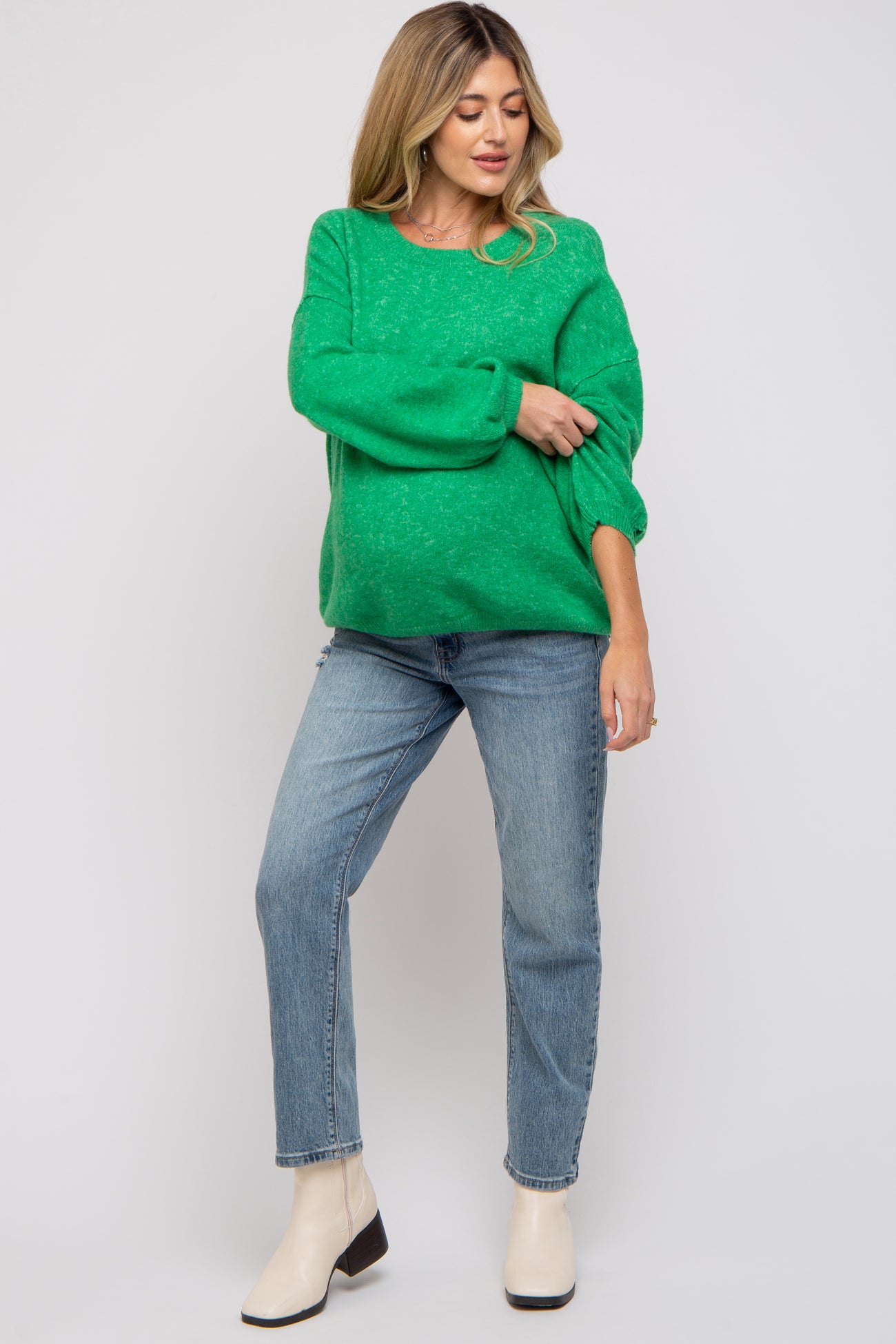 woman wearing a green knit maternity sweater and light blue jeans, standing in a relaxed pose