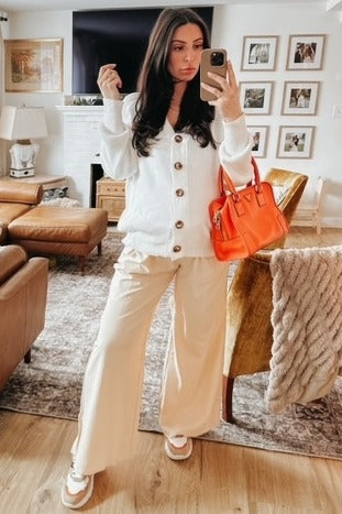 woman wearing a white button-up cardigan and cream wide-leg pants, holding an orange handbag in a cozy living room setting