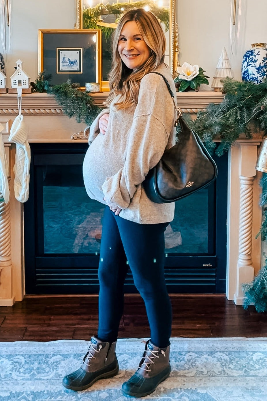 woman wearing a beige oversized maternity sweater and black leggings, holding her baby bump while standing indoors