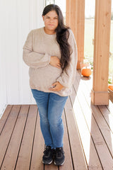 woman wearing a beige maternity sweater and blue jeans, holding her baby bump outdoors on a porch