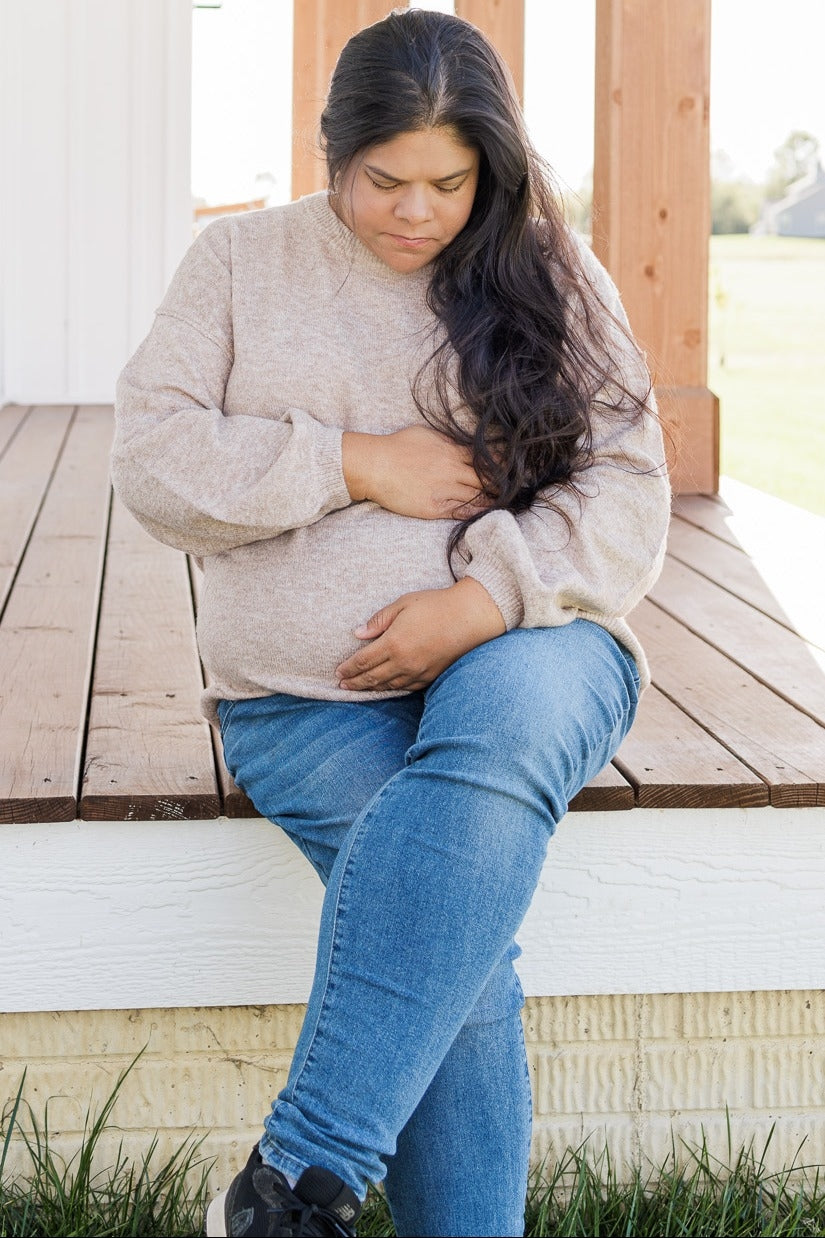 woman wearing a beige oversized maternity sweater and blue jeans, gently cradling her baby bump while sitting on a porch