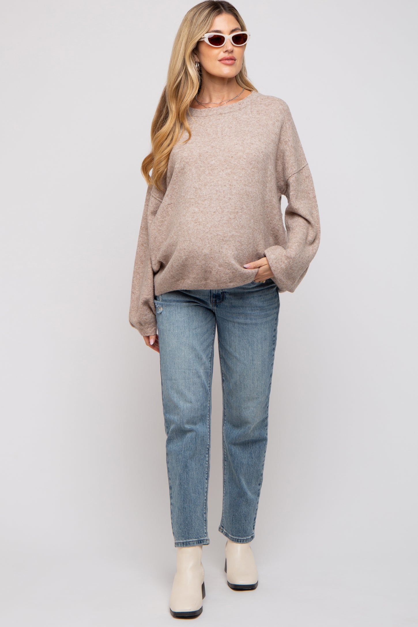 woman wearing a beige oversized maternity sweater and blue jeans, with a visible baby bump and sunglasses
