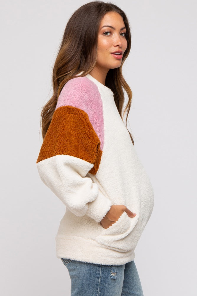 woman wearing a color-blocked cream, pink, orange, and brown maternity sweater with pockets and a visible baby bump