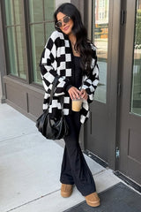 woman wearing a black and white checkered cardigan over a black outfit, holding a drink, standing outside