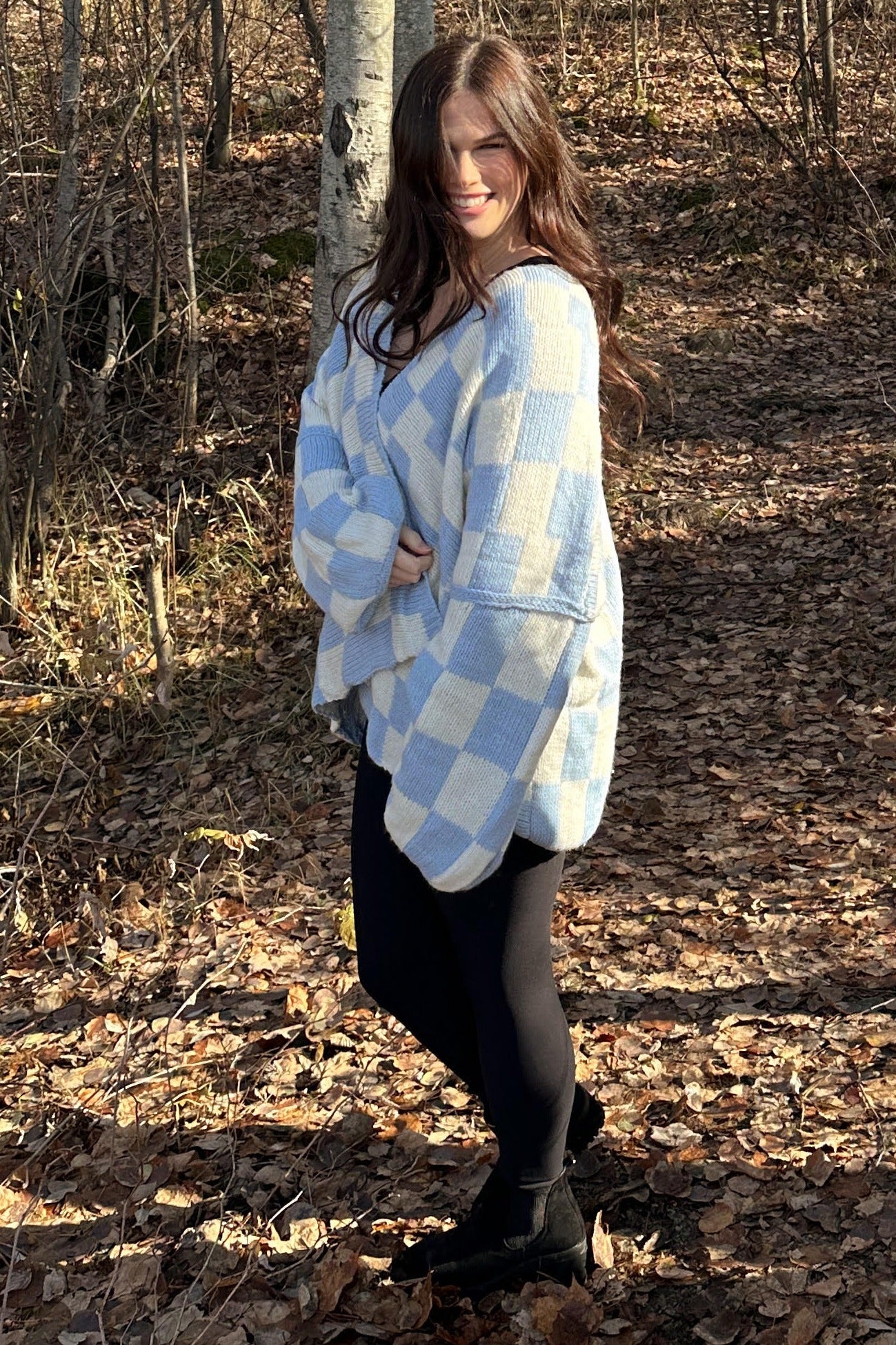 woman wearing a light blue and white checkered oversized sweater with black leggings and boots in a forest setting