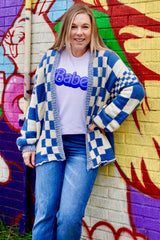 woman wearing a blue and white checkerboard cardigan over a white t-shirt with "Babe" text and blue jeans